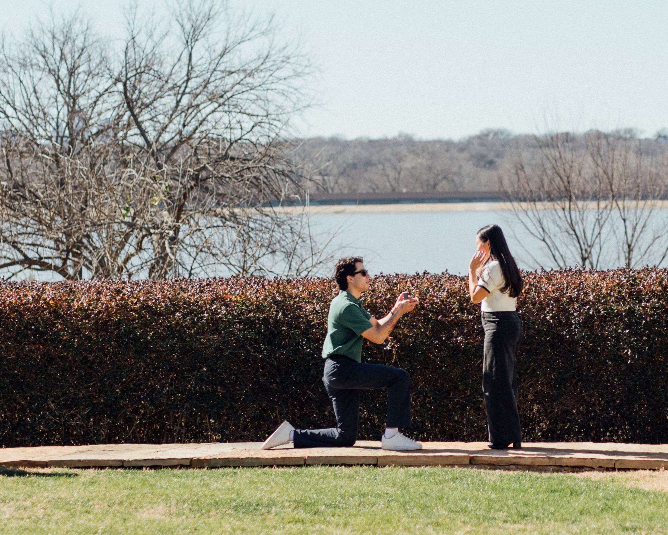 Matthew proposing to Emily on one knee by a lake with Emily covering her mouth in surprise